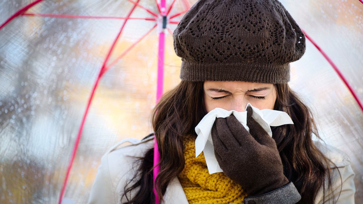 Woman with cold or flu coughing and blowing her nose with a tissue under autumn rain. Brunette female sneezing and wearing warm clothes.