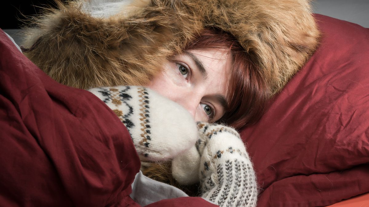 Woman lying in a bed wearing fur hat and mittens and freezing.