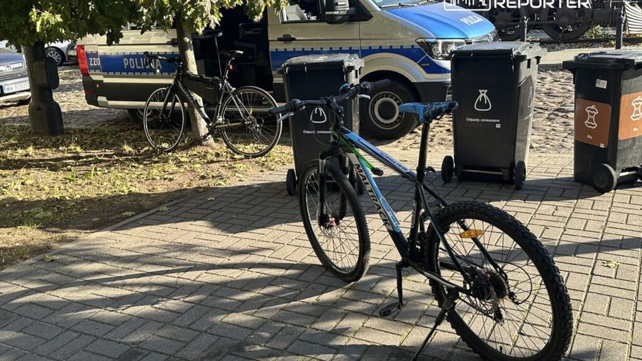 Rower górski zaparkowany obok policyjnego samochodu na ulicy, w tle kontenery na odpady.