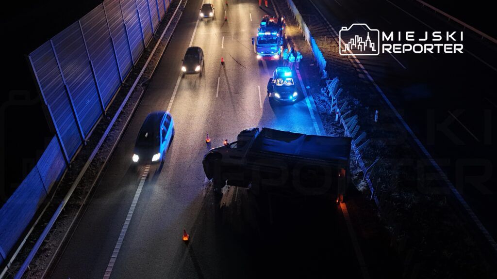 Na drodze leży przewrócony pojazd ciężarowy, a w pobliżu pracują policjanci i strażacy, zabezpieczając miejsce zdarzenia.