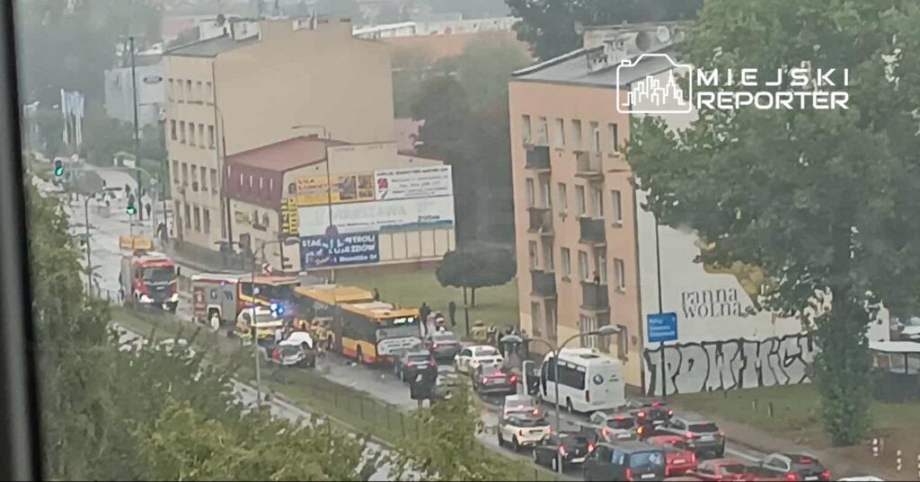 Na ulicy stoją pojazdy ratunkowe, a obok grupka ludzi obserwuje sytuację w deszczowej pogodzie.