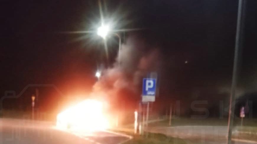 Pojazd płonący na poboczu drogi, otoczony gęstym dymem, w nocy pod oświetleniem ulicznym.