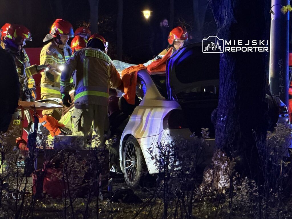 Grupa strażaków w kaskach pracuje nad usunięciem poszkodowanego z wraku samochodu w ciemnym otoczeniu.