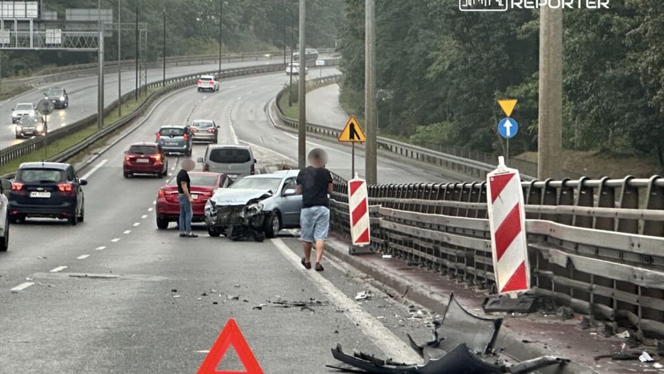 Dwa samochody po kolizji na drodze, mężczyźni sprawdzają uszkodzenia, na jezdni leży rozbita część pojazdu i trójkąt ostrzegawczy.