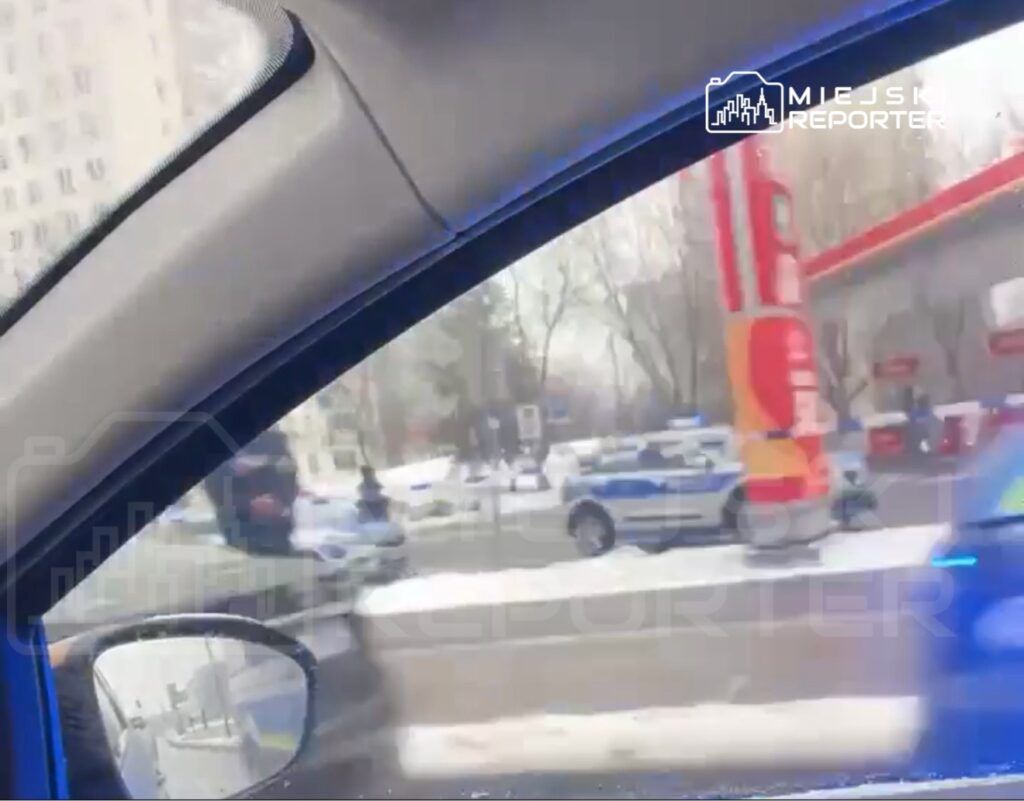 Mężczyzna w czarnej masce stoi obok radiowozu na zaśnieżonej ulicy, w tle widoczne stacje benzynowe.