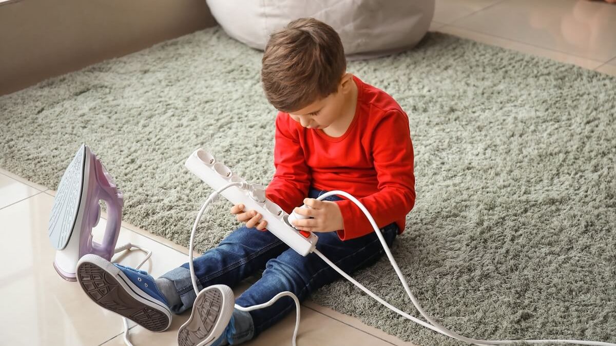 Mały chłopiec podłącza wtyczkę do przedłużacza elektrycznego, siedząc na dywanie obok żelazka