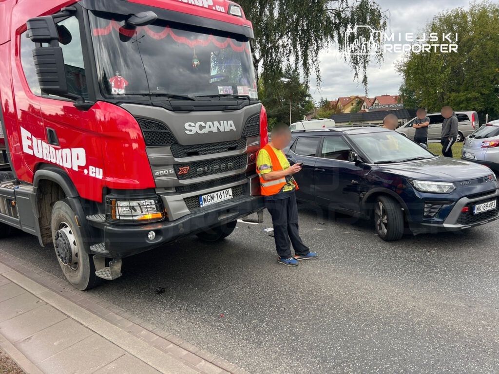 Czerwony ciężarowy samochód marki Scania stoi na drodze, obok niego mężczyzna w odblaskowej kamizelce korzysta z telefonu.