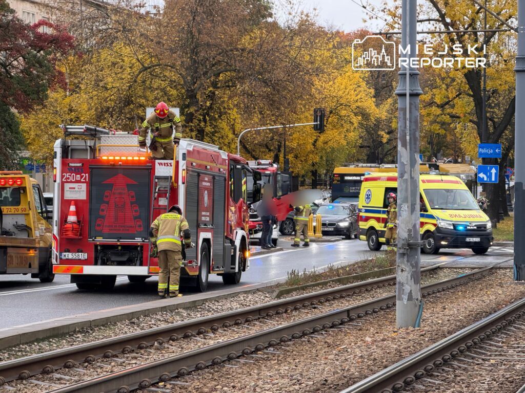 Strażacy z wozu strażackiego rozkładają sprzęt w pobliżu miejsca wypadku na ulicy, obok przejazdu tramwajowego.