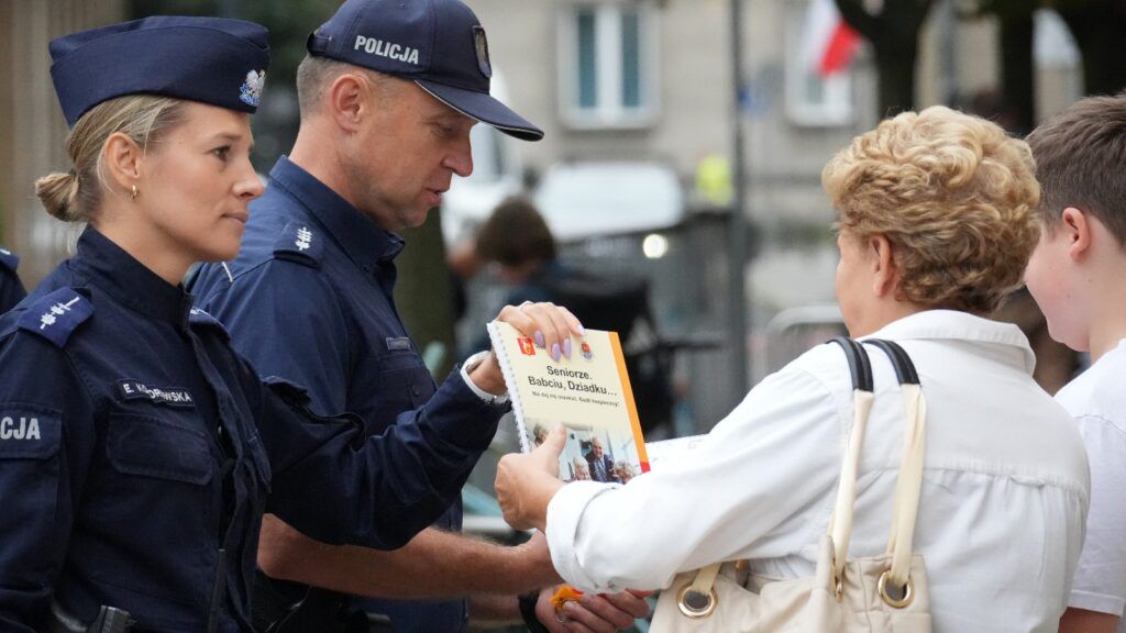 Policjanci wręczają ulotki starszej kobiecie na ulicy, przyciągając uwagę przechodniów.