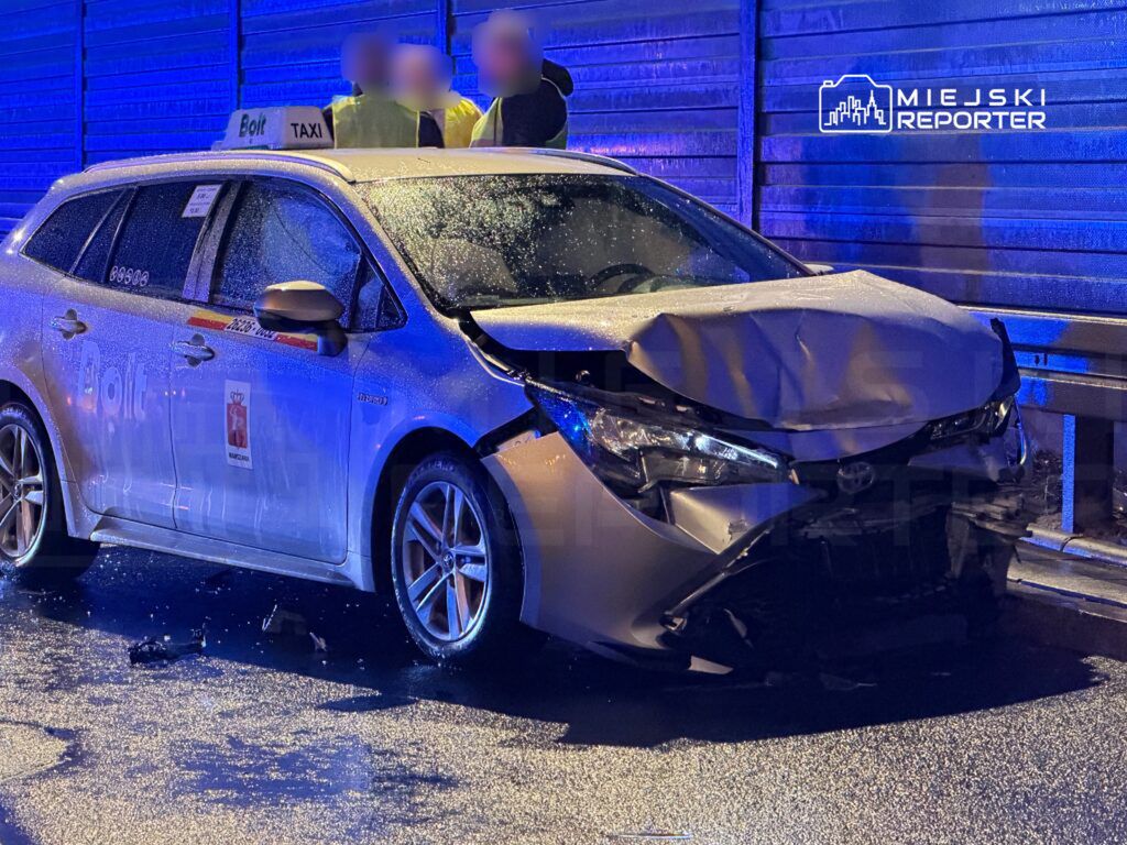 Uszkodzony samochód taxi z widocznym zgniecionym przodem stoi na mokrej nawierzchni, obok grupy osób w kamizelkach odblaskowych.