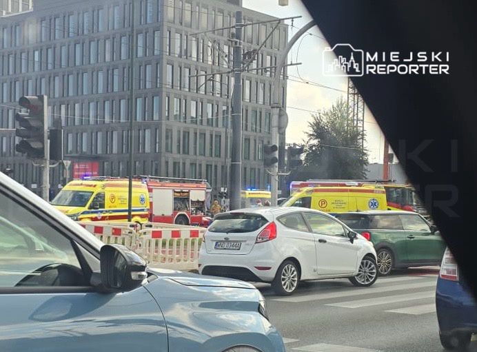 Karetki pogotowia i straż pożarna na miejscu zdarzenia przy nowoczesnym biurowcu, w tle ruch uliczny.