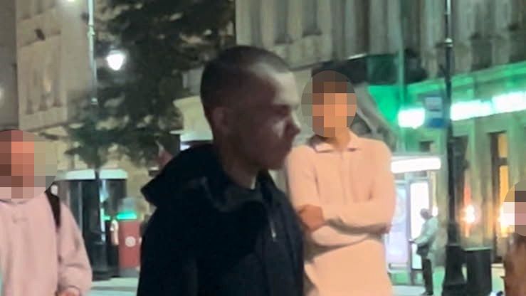 Mężczyzna w czarnej bluzie i krótkich spodenkach spaceruje po ulicy w nocy, obok grupy przechodniów.
