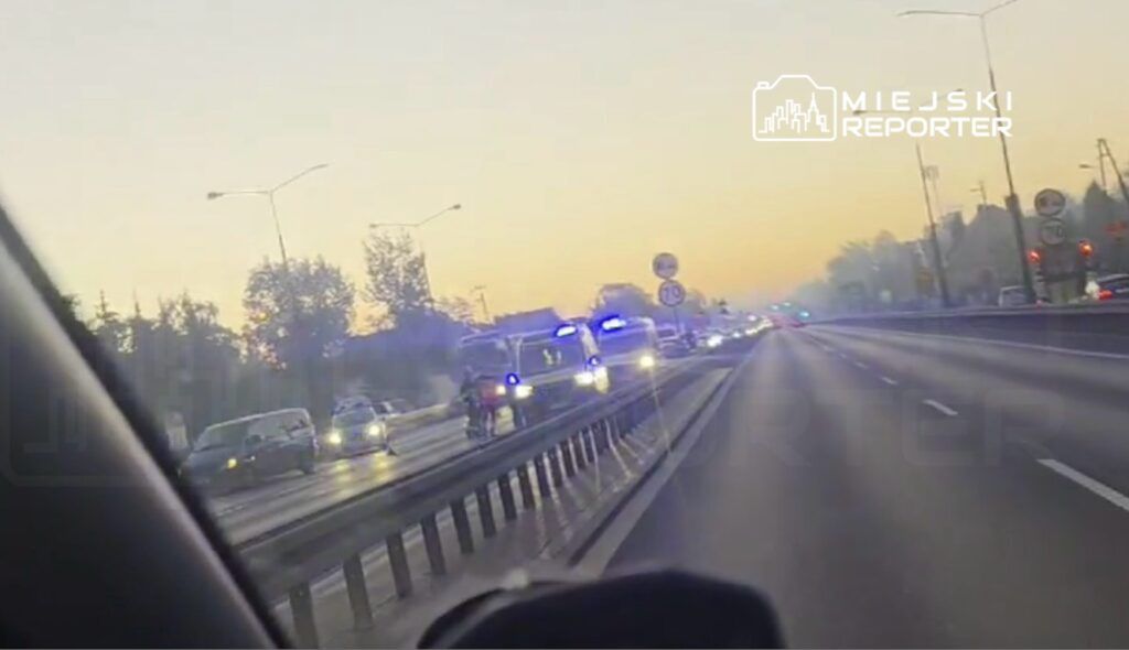 Policyjny radiowóz z włączonymi sygnałami świetlnymi zatrzymuje się na ruchliwej drodze, obok innych pojazdów.