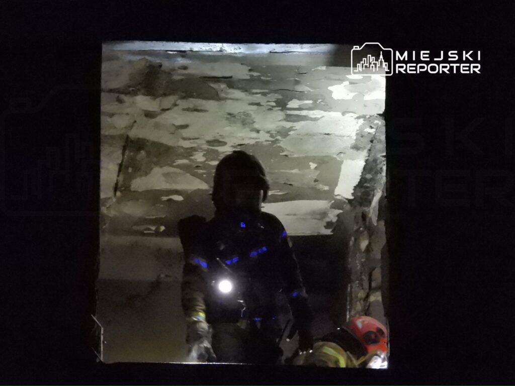 Strażak z latarką przeszukuje ciemne wnętrze budynku, obok niego inny ratownik w hełmie.