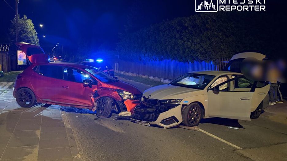 Dwa uszkodzone samochody osobowe zderzone na nocnej ulicy, w tle niebieskie światła radiowozu.