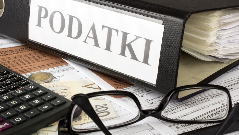Na zdjęciu formularze podatkowe i pieniąze rozrzucone na biurku. Na nich segregator z napisem "podatki"
