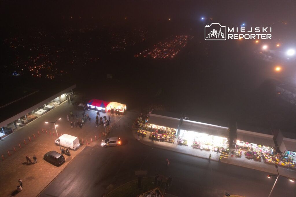 Grupa ludzi zbiera się przy straganach z kwiatami, oświetlonych lampami, na tle nocnego nieba.