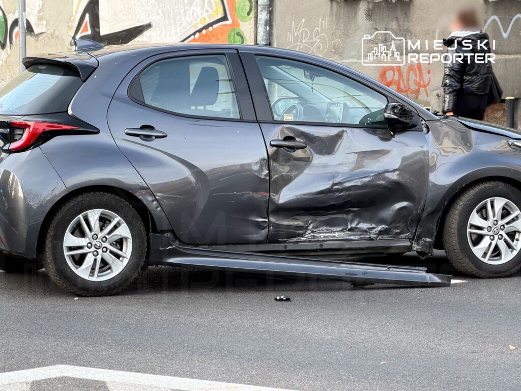 Uszkodzony samochód osobowy w kolorze szarym z wgniecioną boczną częścią zaparkowany na ulicy. W tle widoczna graffiti na murze.
