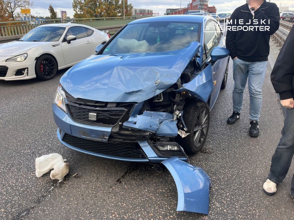 Niebieski samochód z uszkodzoną przednią częścią stoi na drodze, obok dwóch mężczyzn rozmawiających. W tle widoczny biały samochód sportowy.