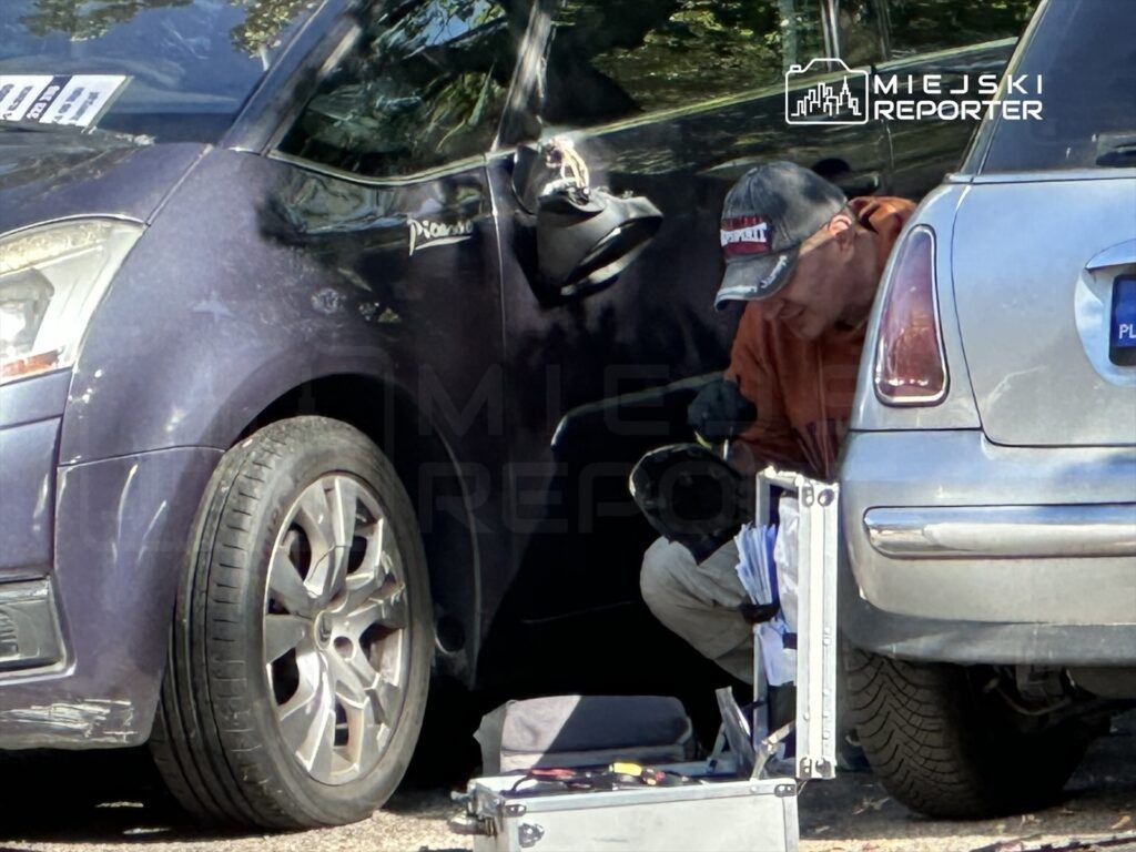 Mężczyzna w czapce naprawia samochód, siedząc na ziemi obok dwóch pojazdów zaparkowanych w słonecznym otoczeniu.