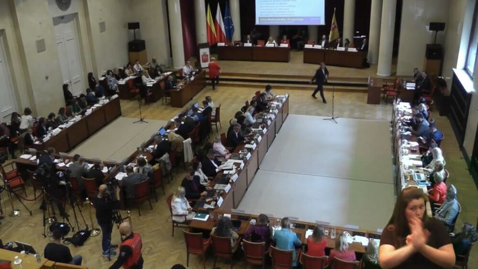 Zgromadzenie osób w sali obrad, niektórzy uczestnicy siedzą przy stołach, na ekranie prezentacja.