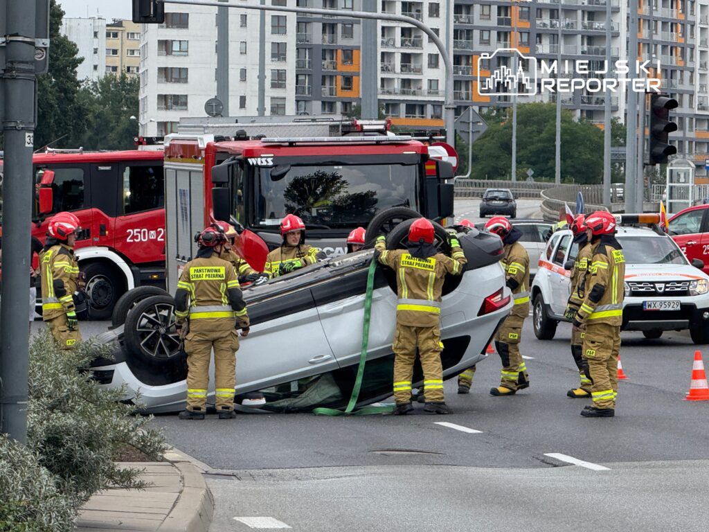 Grupa strażaków w mundurach próbuje postawić na koła przewrócone auto na ruchliwej drodze.