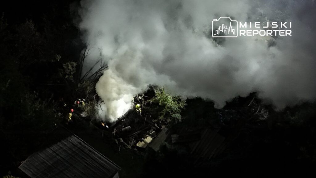 Strażacy gaszą pożar w zniszczonym budynku, otoczonym gęstym dymem i roślinnością, w nocy.