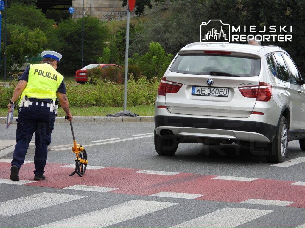 Policjant w kamizelce odblaskowej mierzy odległość na przejściu dla pieszych, obok zatrzymany samochód marki BMW.