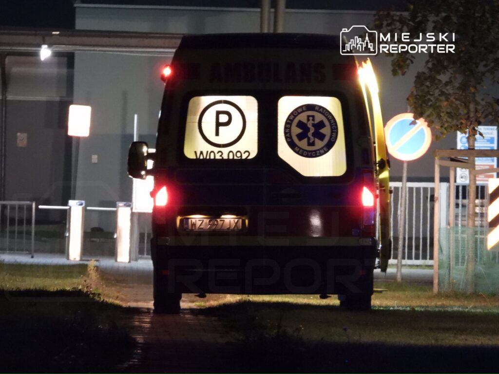 Karetka pogotowia z włączonymi światłami parkowania stoi na chodniku nocą, w tle widoczne znaki drogowe.