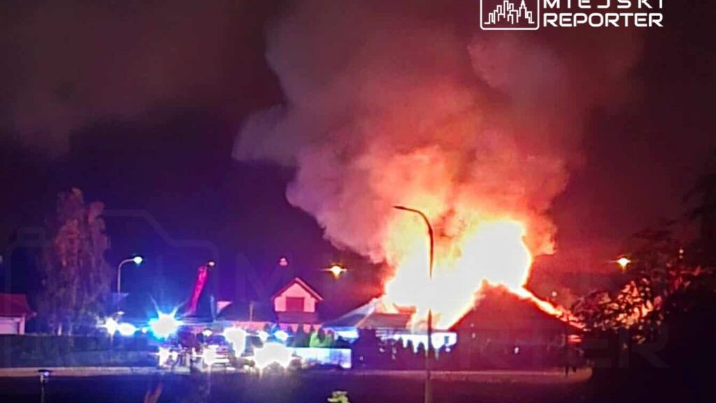 Płonący budynek w nocy, otoczony przez straż pożarną i pojazdy ratunkowe, z intensywnym ogniem i dymem.