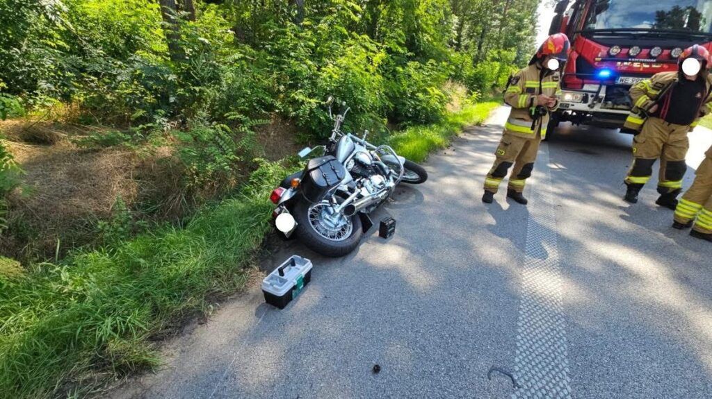 Motocykl leży na boku przy drodze, a obok stoją strażacy w mundurach ratunkowych.
