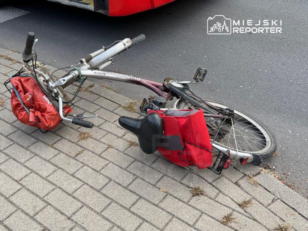 Złamany rower leży na chodniku, z czerwoną torbą przymocowaną do kierownicy i widoczną tylną lampką.