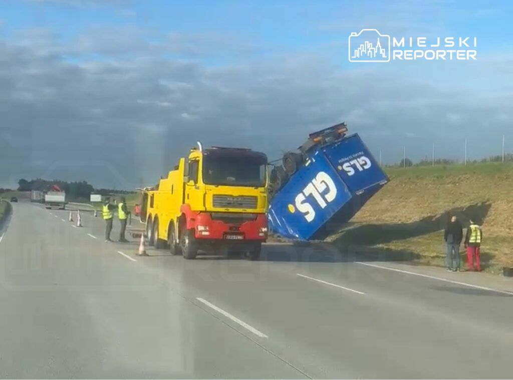 Na drodze leży przewrócona ciężarówka z niebieską naczepą, obok dwóch mężczyzn w kamizelkach odblaskowych.