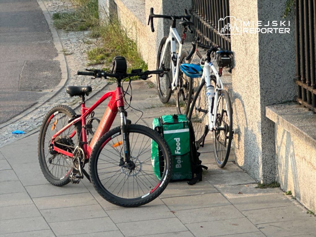 Dwa rowery, jeden czerwony górski, drugi biały szosowy, stoją obok torby dostawczej na chodniku.