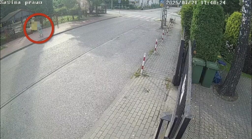 Dziecko w kolorowych spodniach stoi na chodniku obok ogrodzenia, w tle widoczna ulica i zieleń.