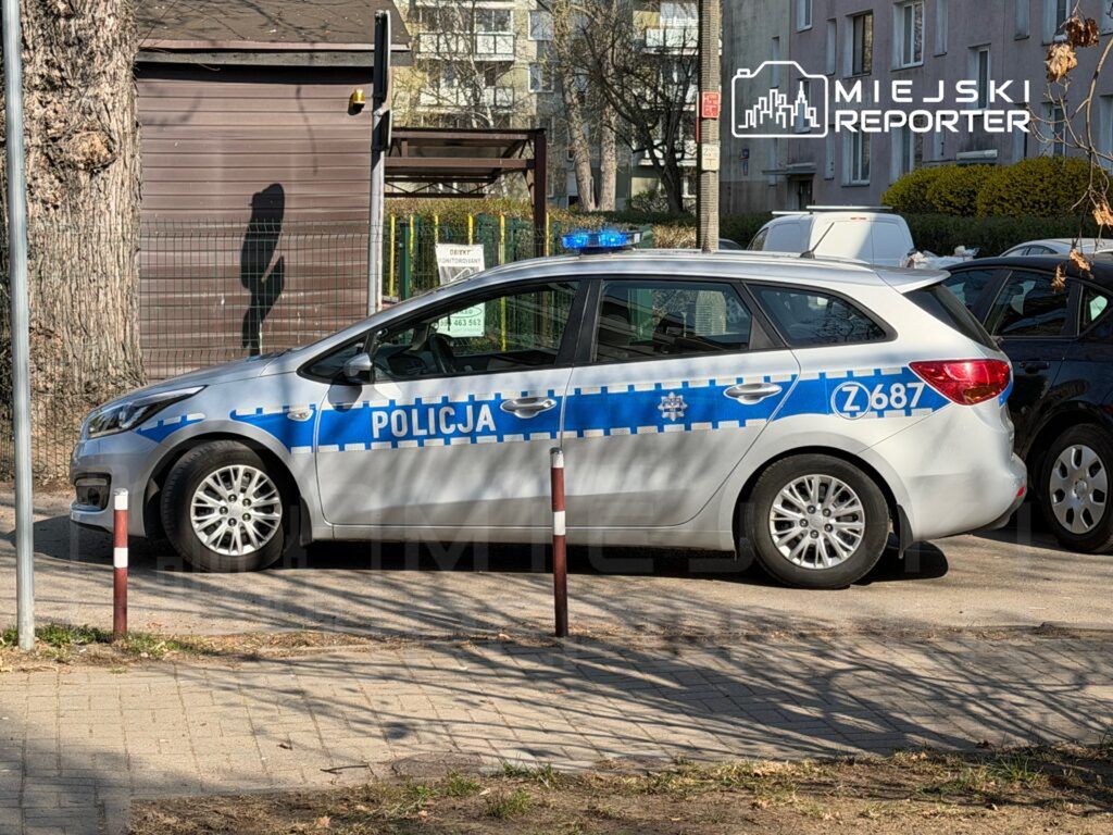 Policyjny radiowóz zaparkowany na osiedlowej ulicy, obok ogrodzenia z zielonym tłem.