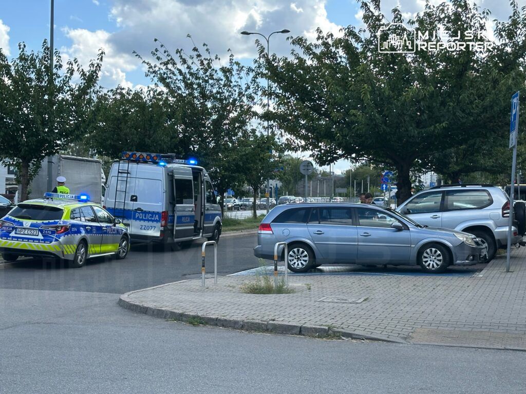 Policyjny radiowóz oraz furgonetka z włączonymi sygnałami świetlnymi stoją obok uszkodzonego samochodu na parkingu.