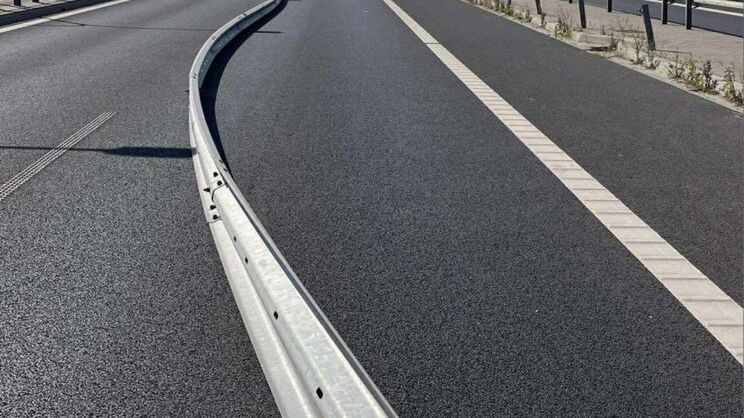 Zakrzywiona bariera ochronna na szerokiej, asfaltowej jezdni, w tle oznakowanie drogi i pojazdy.