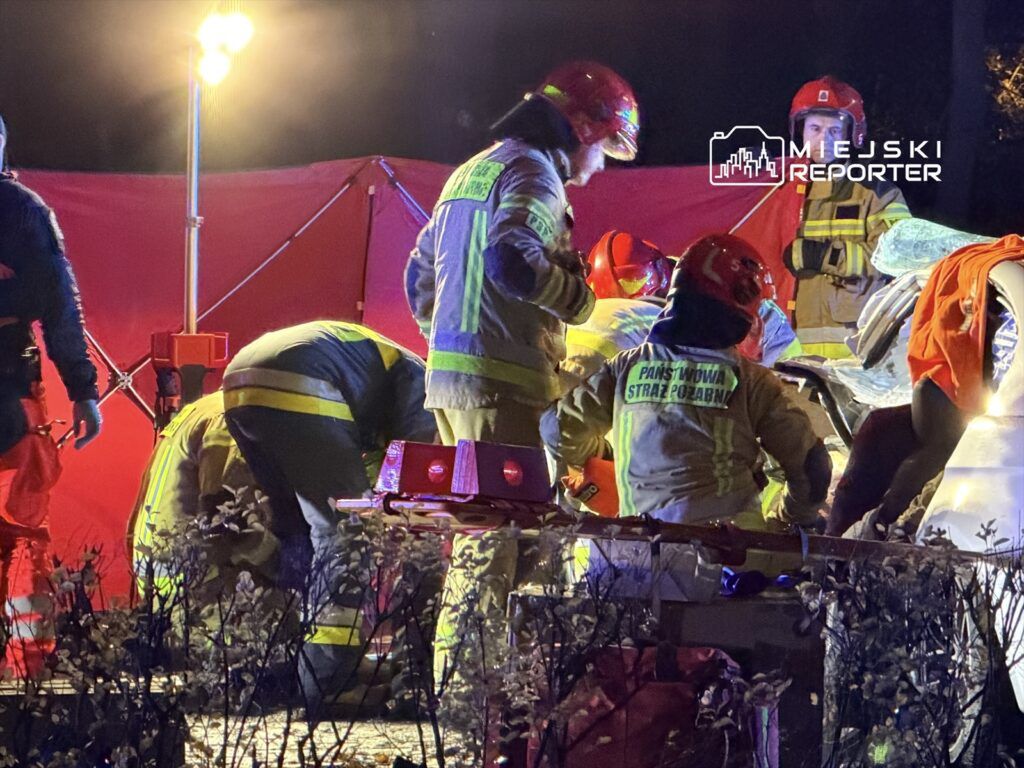 Grupa strażaków w mundurach ratunkowych pracuje przy wypadku samochodowym, otoczeni przez światła i czerwony namiot.