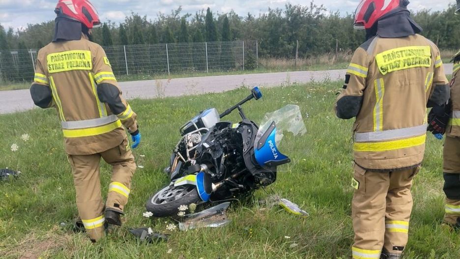 Strażacy w hełmach oceniają przewrócony motocykl na poboczu drogi, w tle zieleń i ogrodzenie.
