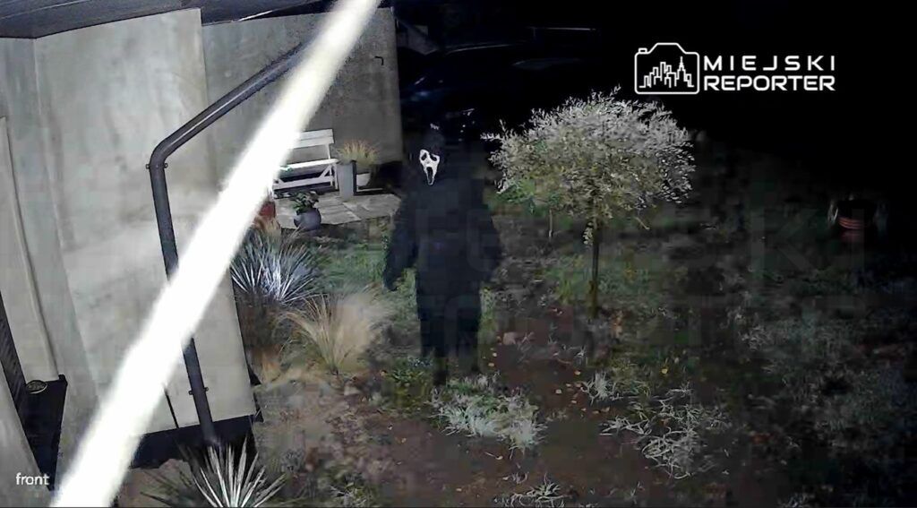 Osoba w czarnym stroju z maską stoi na podjeździe, otoczona przez roślinność w nocy.