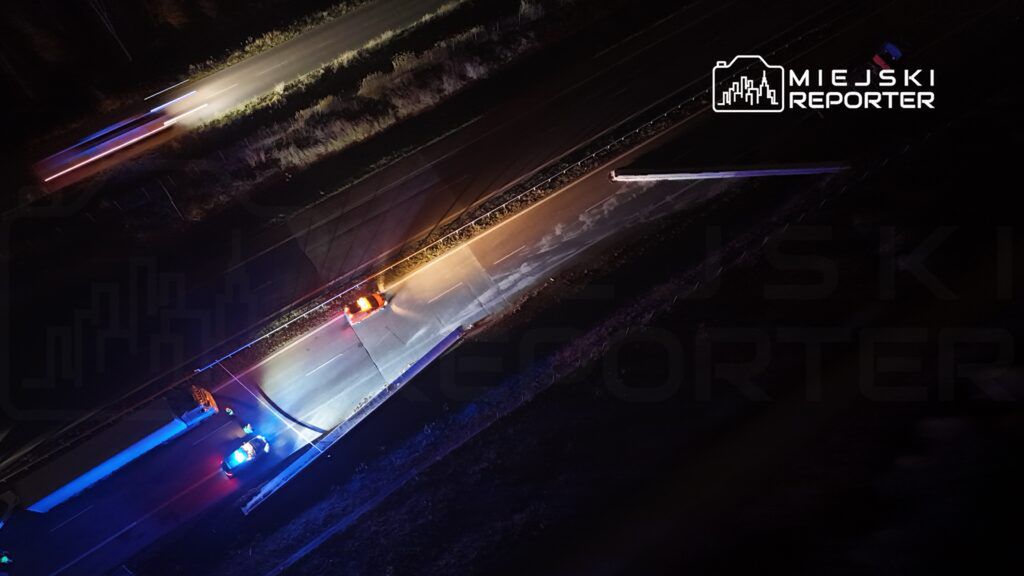 Nocna scena na drodze z policyjnymi pojazdami włączonymi sygnałami świetlnymi, w tle ciemny las.