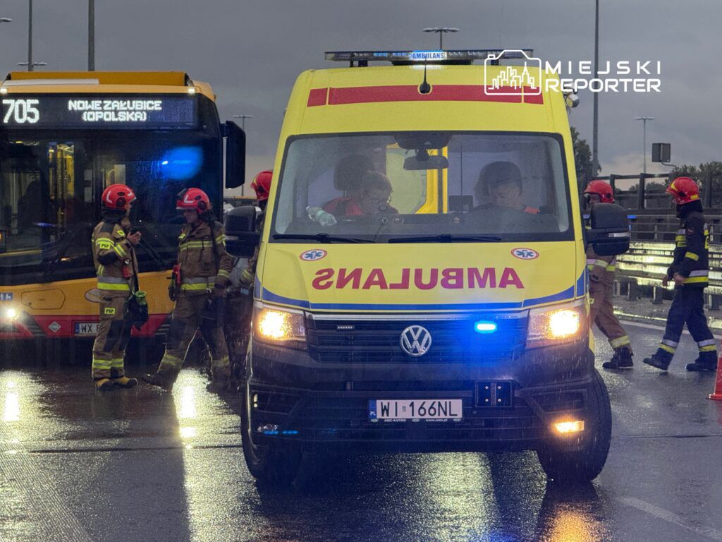 Zespół ratowników w hełmach omawia sytuację obok karetki na mokrej drodze, a w tle widać autobus.
