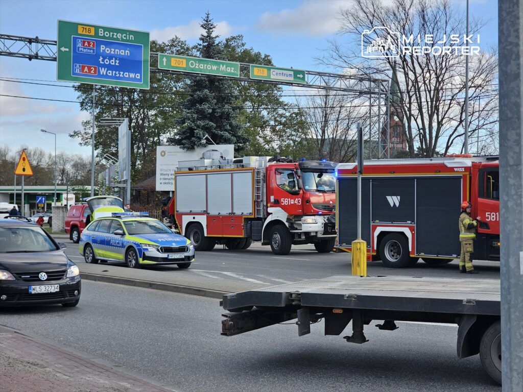 Na skrzyżowaniu ulicy stoi wóz strażacki oraz radiowóz policji, a w tle widoczne znaki drogowe kierujące do Poznania i Warszawy.
