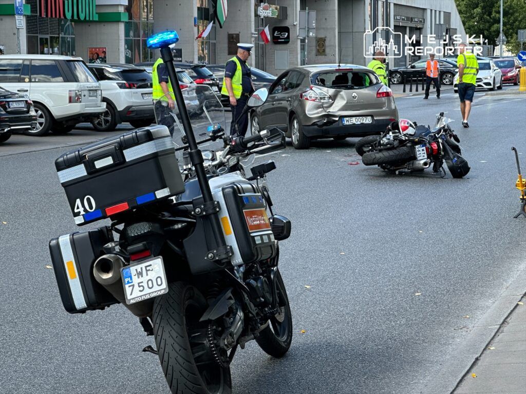 Motocykl leży na jezdni obok uszkodzonego samochodu, a policjanci w kamizelkach odblaskowych zabezpieczają miejsce wypadku.