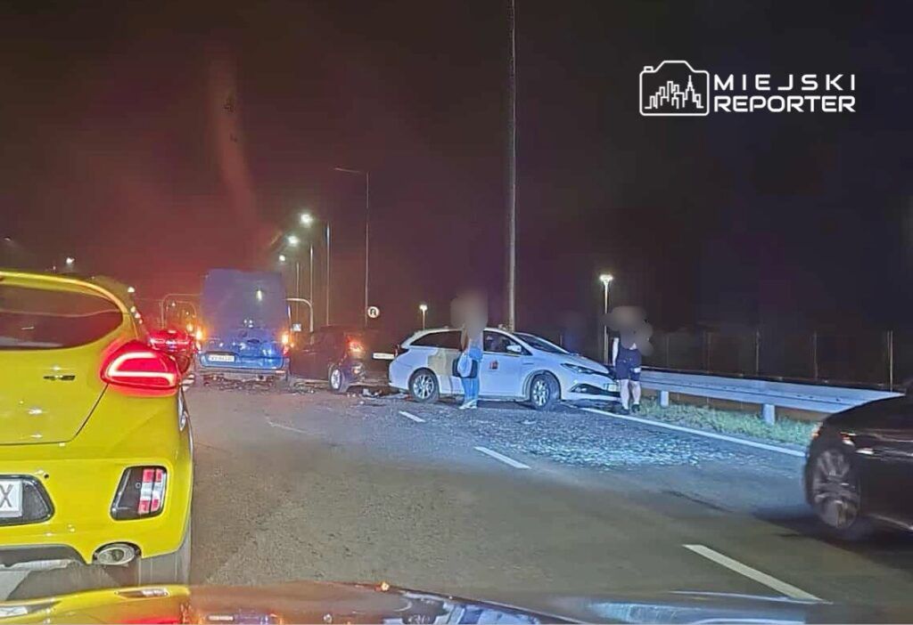 Zdarzenie drogowe z udziałem kilku pojazdów na oświetlonej ulicy w nocy, z osobami sprawdzającymi uszkodzenia.