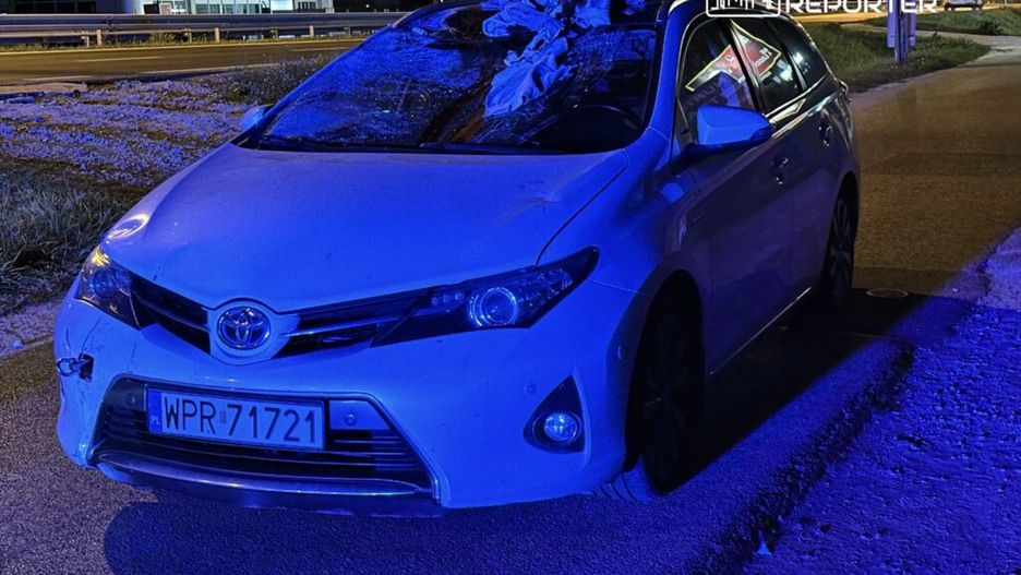 Uszkodzony biały samochód osobowy marki Toyota zaparkowany na poboczu drogi, oświetlony niebieskimi światłami.