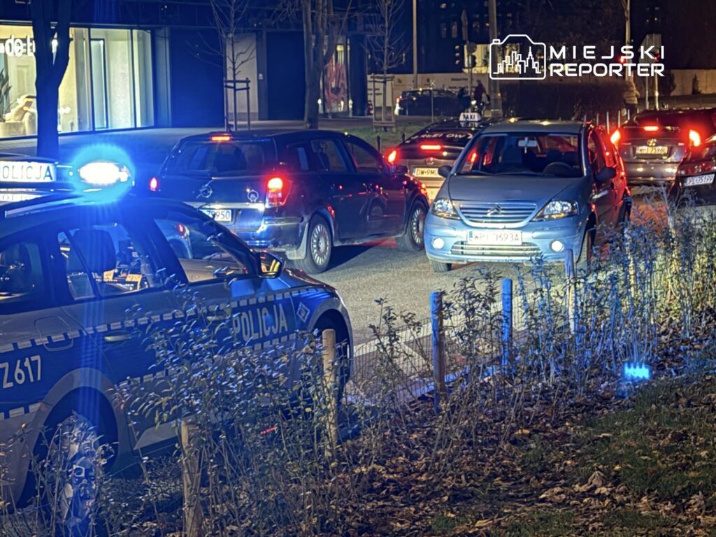 Policyjny radiowóz z włączonymi sygnałami świetlnymi zatrzymuje ruch na ulicy, obok stoją samochody osobowe.