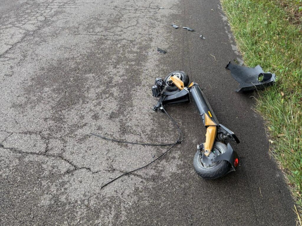 Złamana hulajnoga leży na asfaltowej drodze, obok widoczne fragmenty plastikowych osłon.