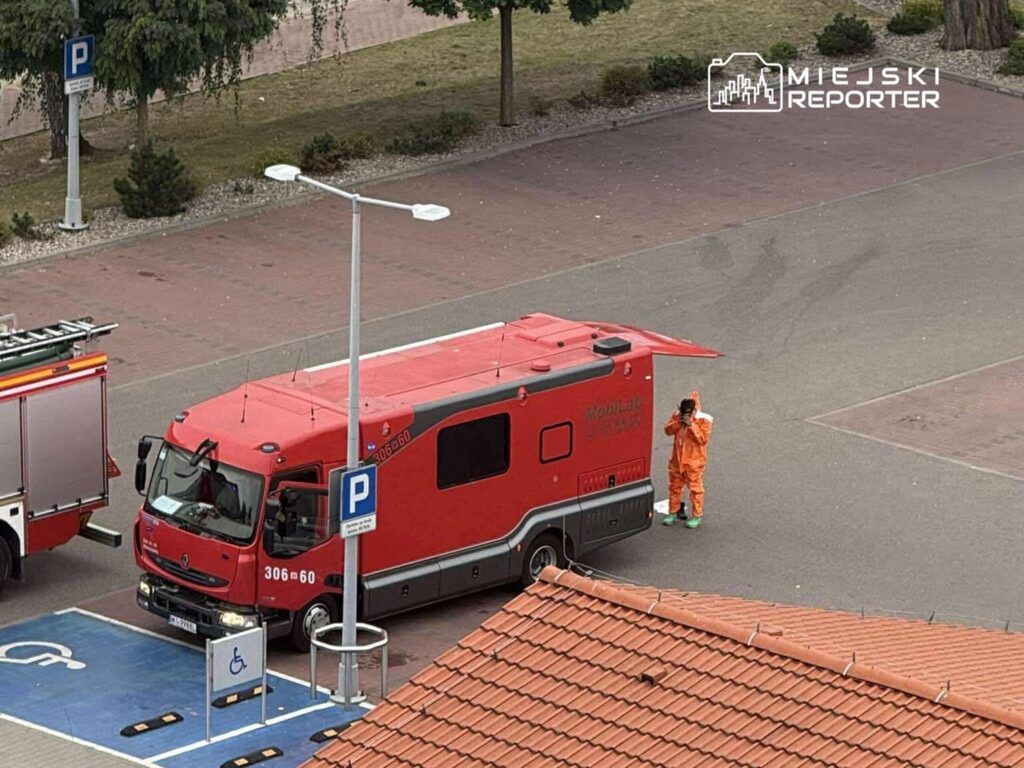 Czerwony pojazd ratunkowy parkowany na placu, a obok stoi osoba w pomarańczowym kombinezonie, trzymająca aparat.